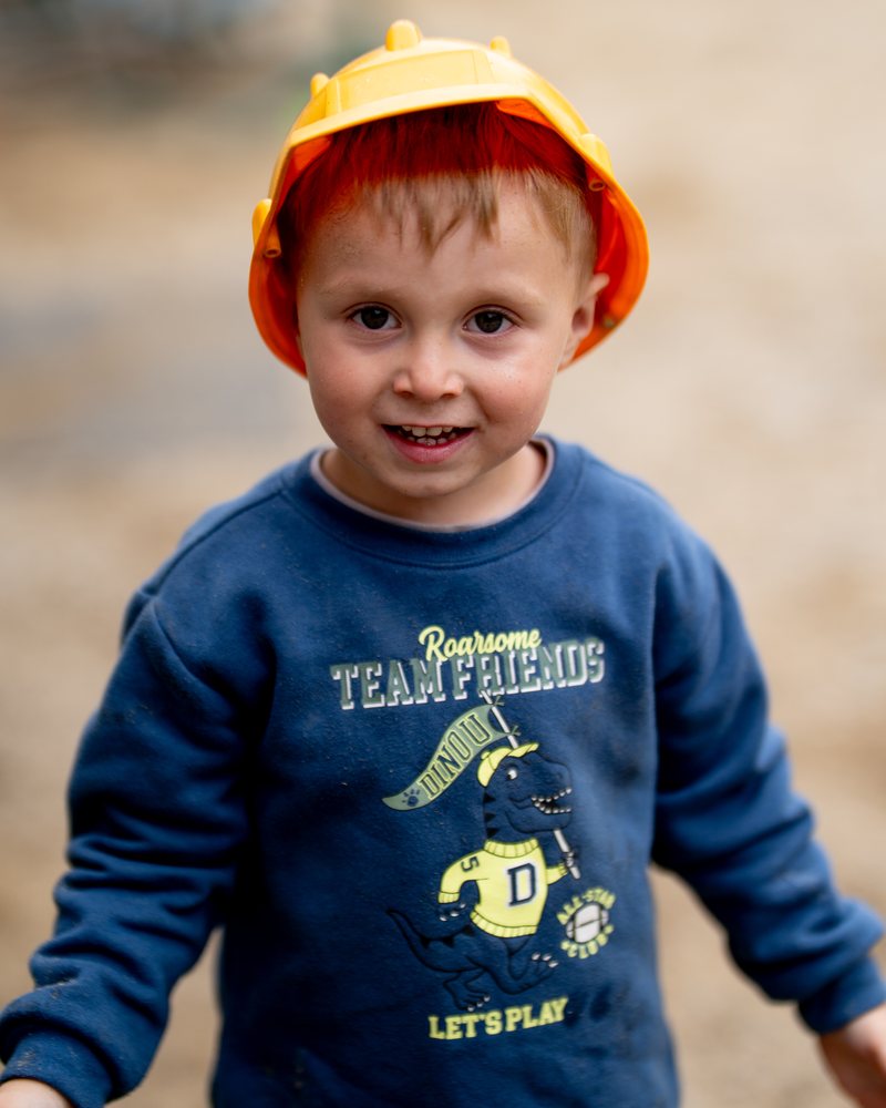 A boy in a yellow toy hard hat smiling on the playground