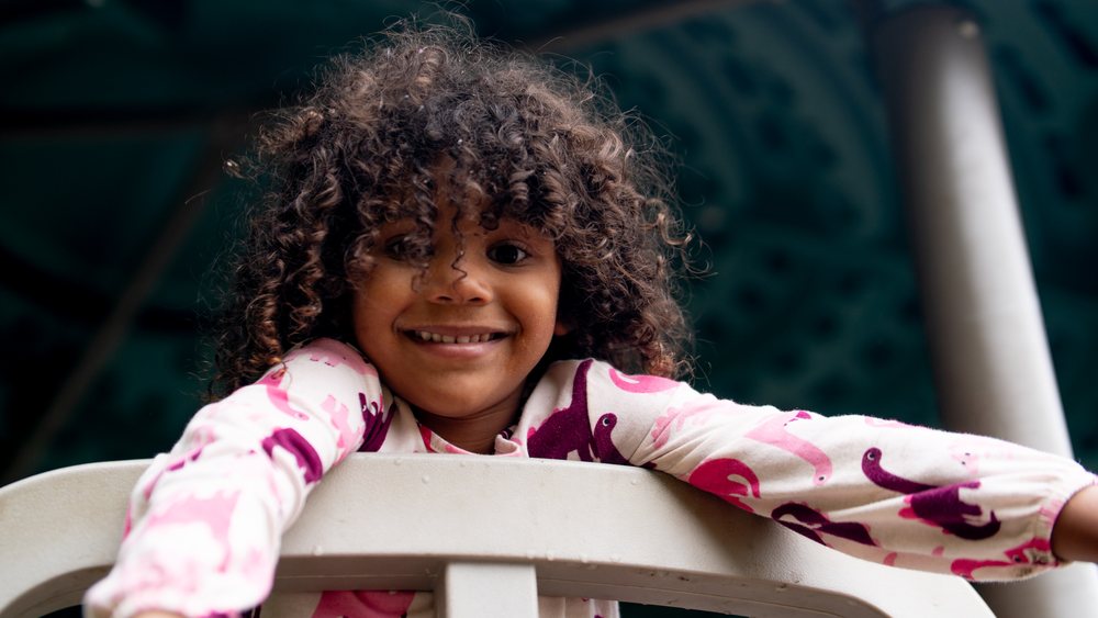 A curly-haired girl in a pink top smiling outdoors