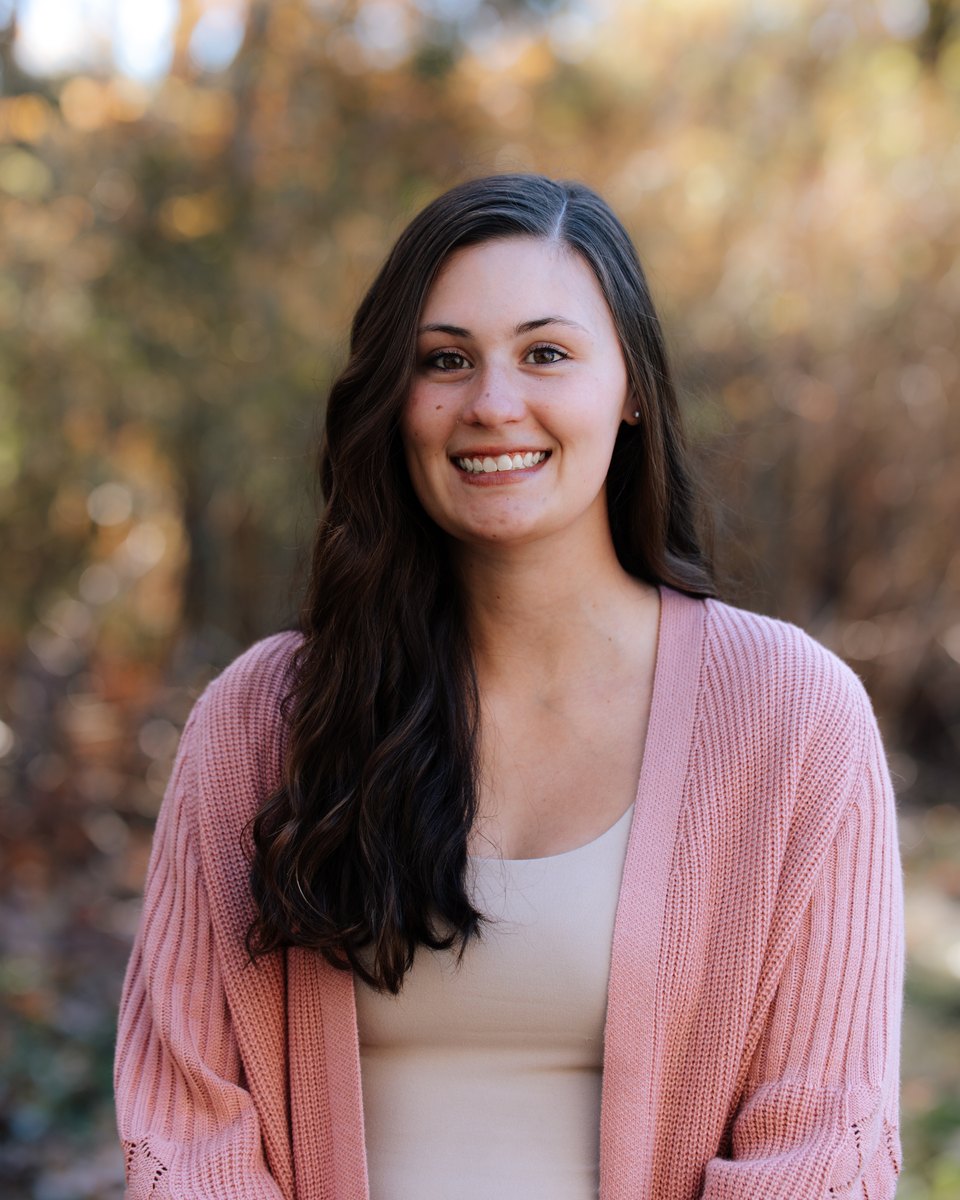 Alexis smiling outdoors in a pink cardigan, autumn trees behind her