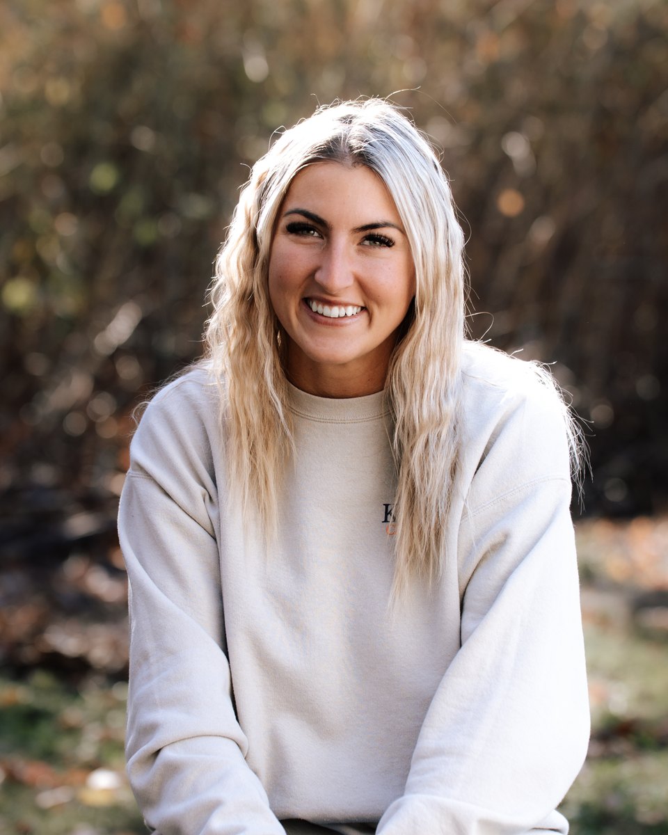 Eliza smiling outdoors in a cream sweatshirt, autumn trees behind her