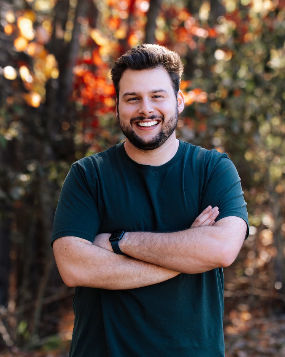 Ganan smiling with his arms crossed in front of autumn leaves