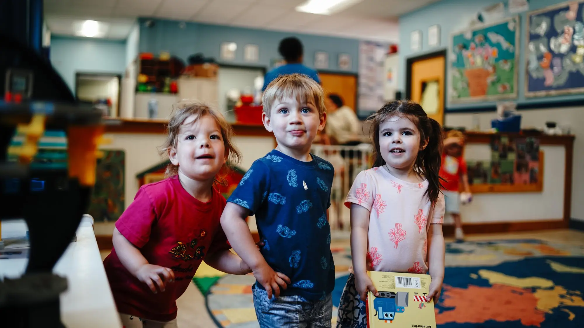Three children leaning in together in a colourful Kids & Co. classroom
